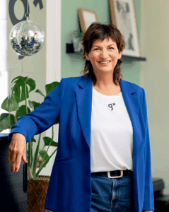 Eine l&auml;chelnde Frau mit kurzen braunen Haaren, die zum Team der werbeagentur Karlsruhe geh&ouml;rt, tr&auml;gt einen blauen Blazer &uuml;ber einem wei&szlig;en T-Shirt und Jeans. Sie steht in einem Innenraum in der N&auml;he einer Pflanze und einer verspiegelten Discokugel, mit gerahmter Kunst an der gr&uuml;nen Wand hinter ihr.