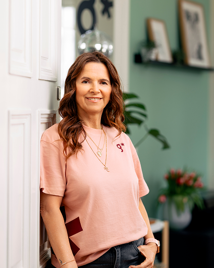 Eine Frau mit gewellten braunen Haaren steht l&auml;chelnd vor einer wei&szlig;en T&uuml;r in einem gem&uuml;tlichen Raum mit gr&uuml;nen W&auml;nden, gerahmten Kunstwerken und Pflanzen - ihr rosa T-Shirt mit dem Symbol der Werbeagentur Karlsruhe und die mehrlagigen goldenen Halsketten vervollst&auml;ndigen ihr stilvolles Aussehen.