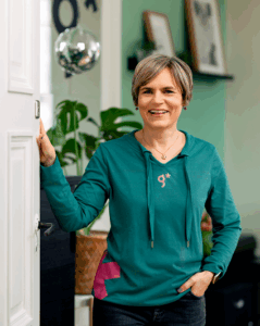 Eine Frau mit kurzen Haaren und einem blaugr&uuml;nen Kapuzenpullover l&auml;chelt, w&auml;hrend sie in einem Raum mit gr&uuml;nen W&auml;nden, Zimmerpflanzen und gerahmten Bildern steht - ein kreativer Raum, der die Atmosph&auml;re einer werbeagentur Karlsruhe widerspiegelt. Sie st&uuml;tzt sich mit einer Hand auf einen wei&szlig;en T&uuml;rrahmen.