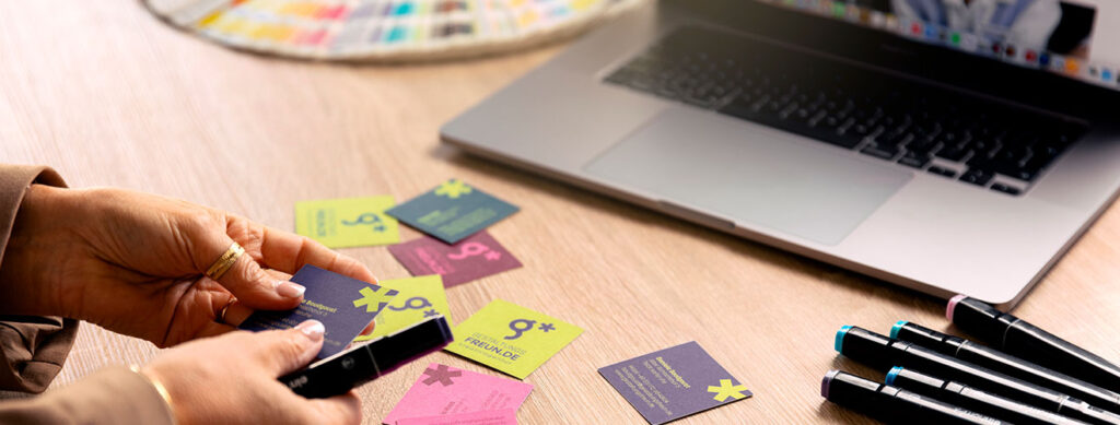 home_hand_vika_faecher Eine Person ordnet bunte Visitenkarten auf einem Schreibtisch neben einem Laptop, Markern und einem Farbmusterbuch an - perfekt, um sich in einer werbeagentur Karlsruhe inspirieren zu lassen.