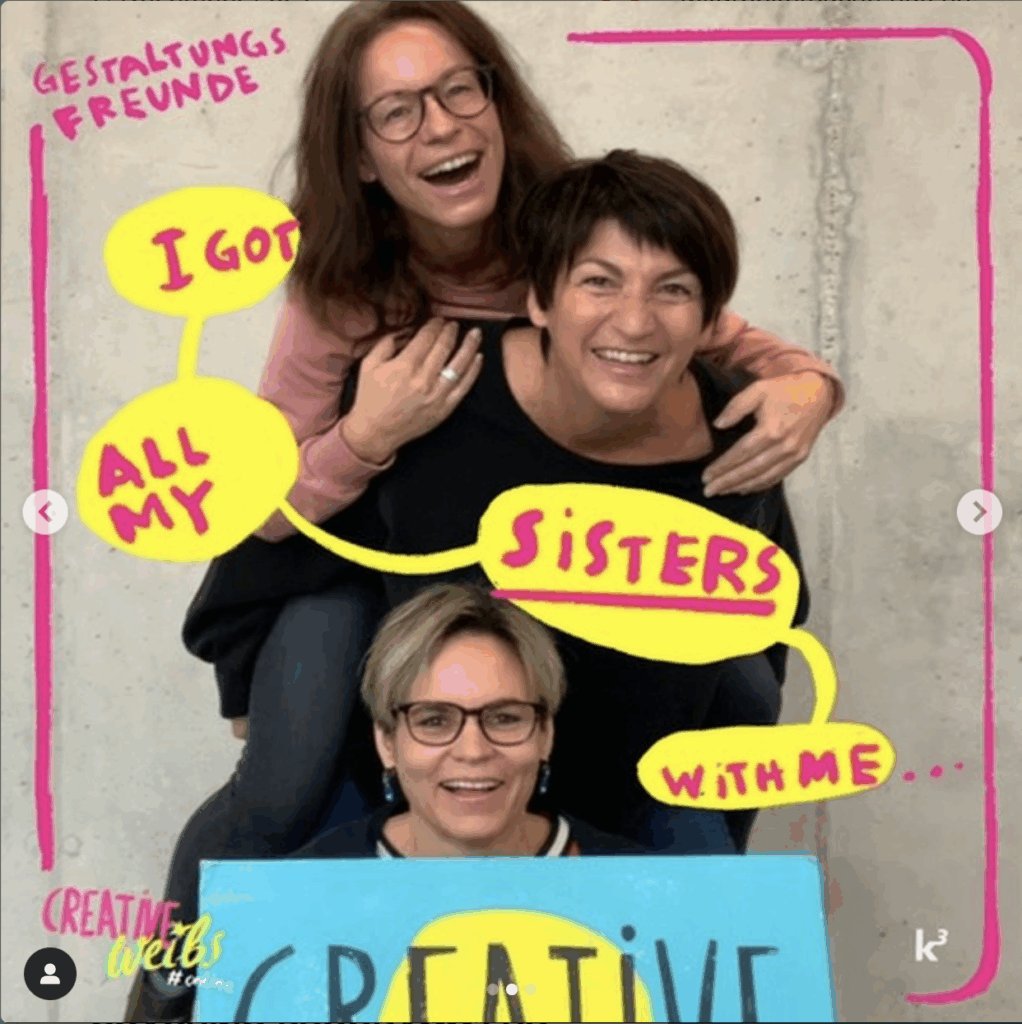 Drei l&auml;chelnde Frauen, die in spielerischer Pose &uuml;bereinander gestapelt sind, teilen sich Sprechblasen im Comic-Stil mit der Aufschrift "I got all my sisters with me..." - ein lebhafter Team-Moment in einer werbeagentur Karlsruhe, wobei eine stolz ein "CREATIVE"-Schild h&auml;lt.
